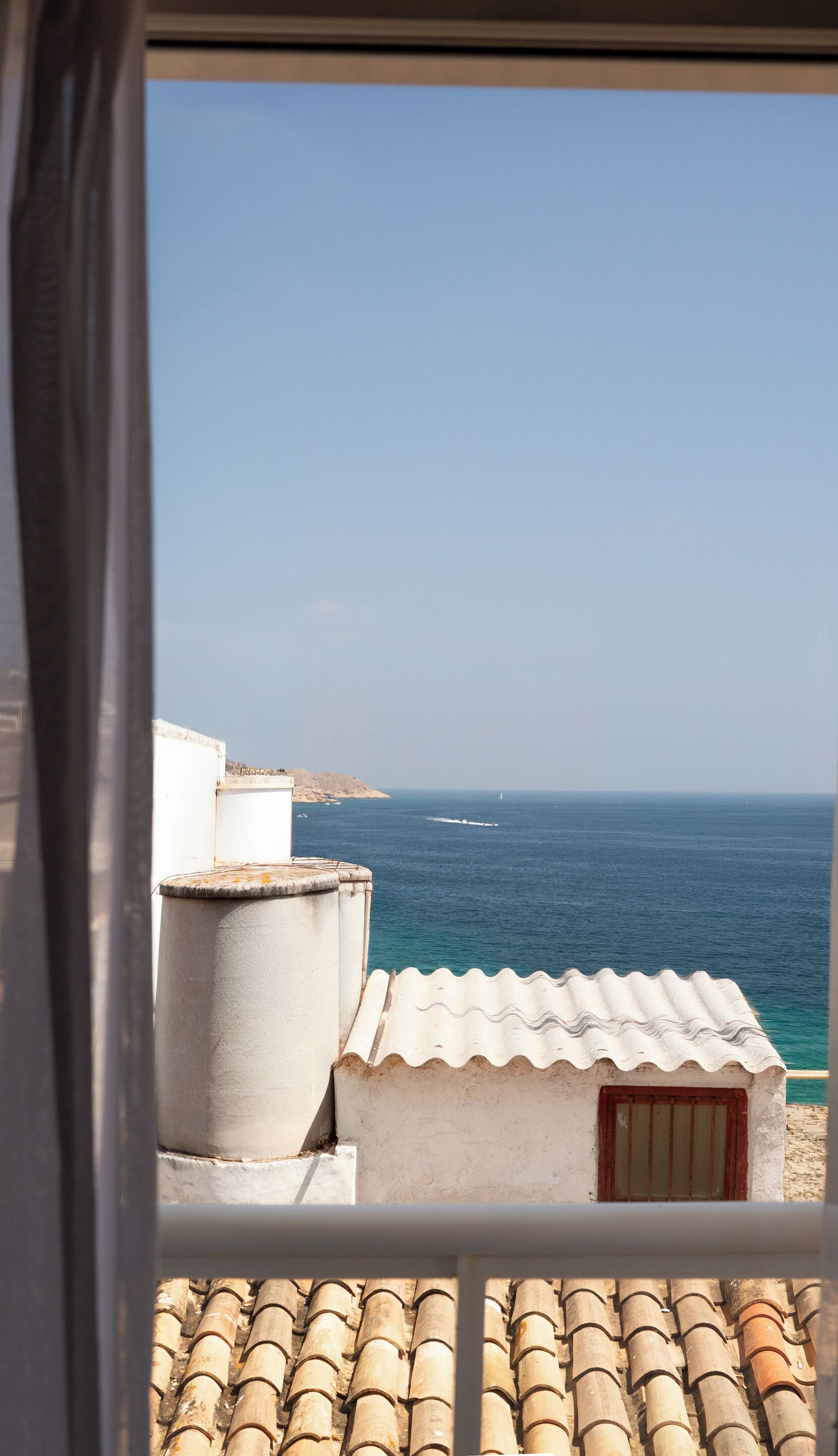 Une vue sur la côte depuis un balcon, avec un ciel dégagé, un voilier au loin et une côte rocheuse.