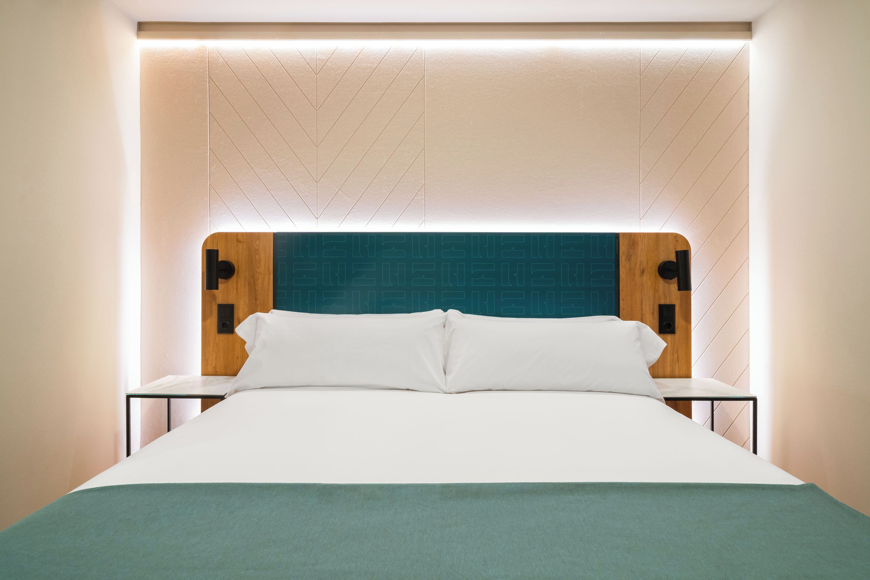 A modern hotel room with a bed, nightstands, and a wooden headboard with a teal accent.