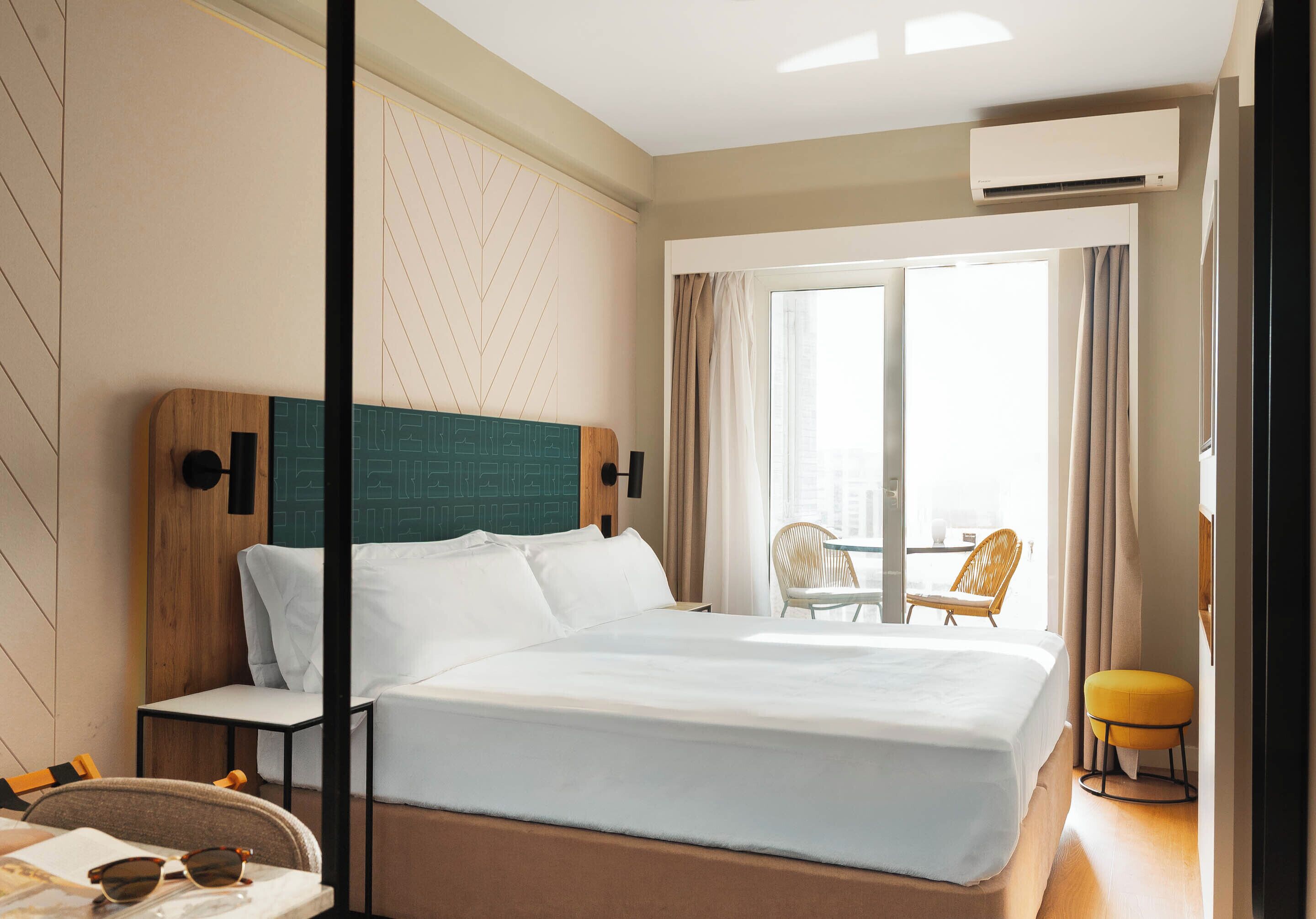 Une chambre d’hôtel comprenant un lit, une table de chevet, une lampe, une chaise, une petite table et une fenêtre avec des rideaux.