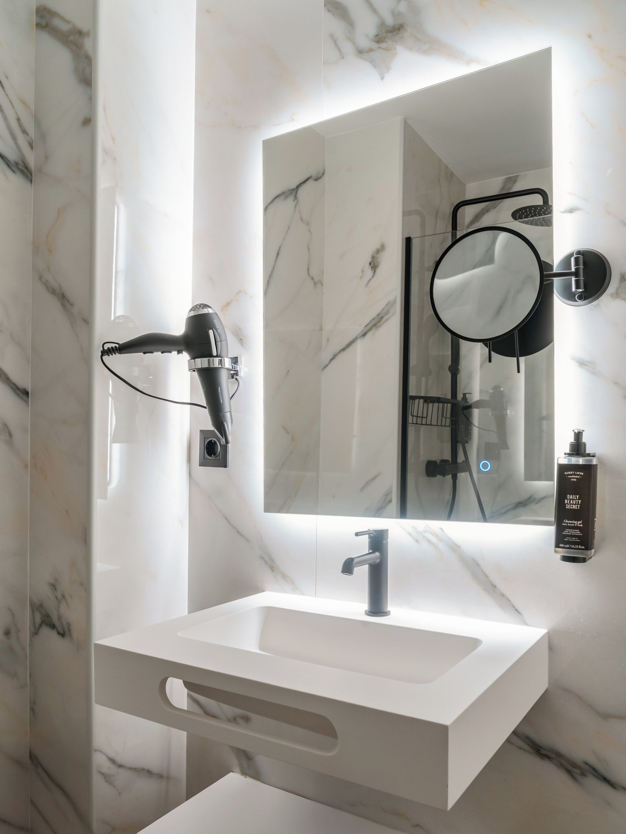 A bathroom with a white marble sink, a handheld hair dryer, a round mirror, and a bottle of shampoo.