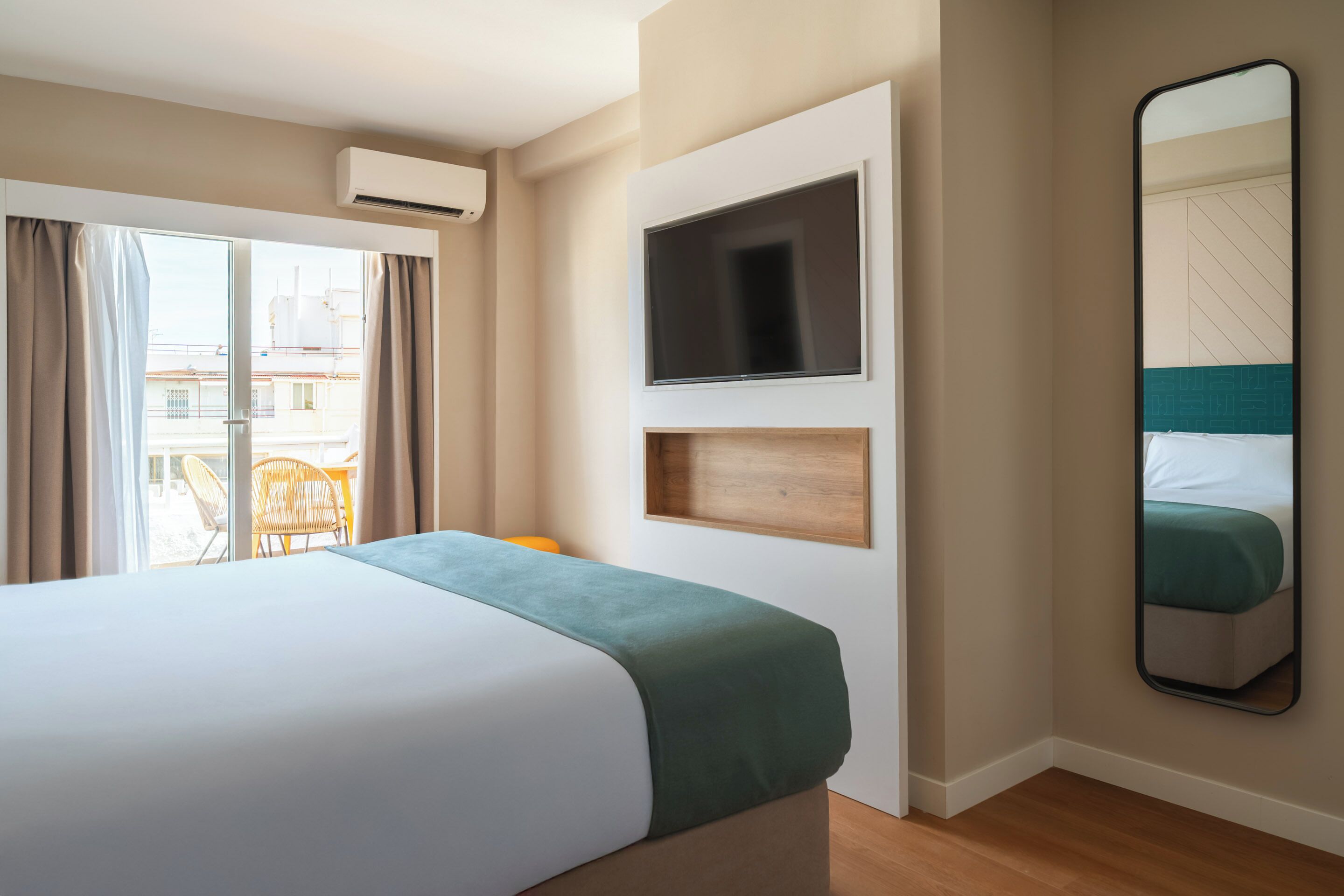 A hotel room with a bed, a TV, a mirror, and a window with curtains.