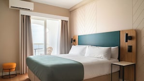 Una habitación de hotel moderna con una cama grande, un cabecero de madera, una mesita auxiliar pequeña y vistas al exterior.