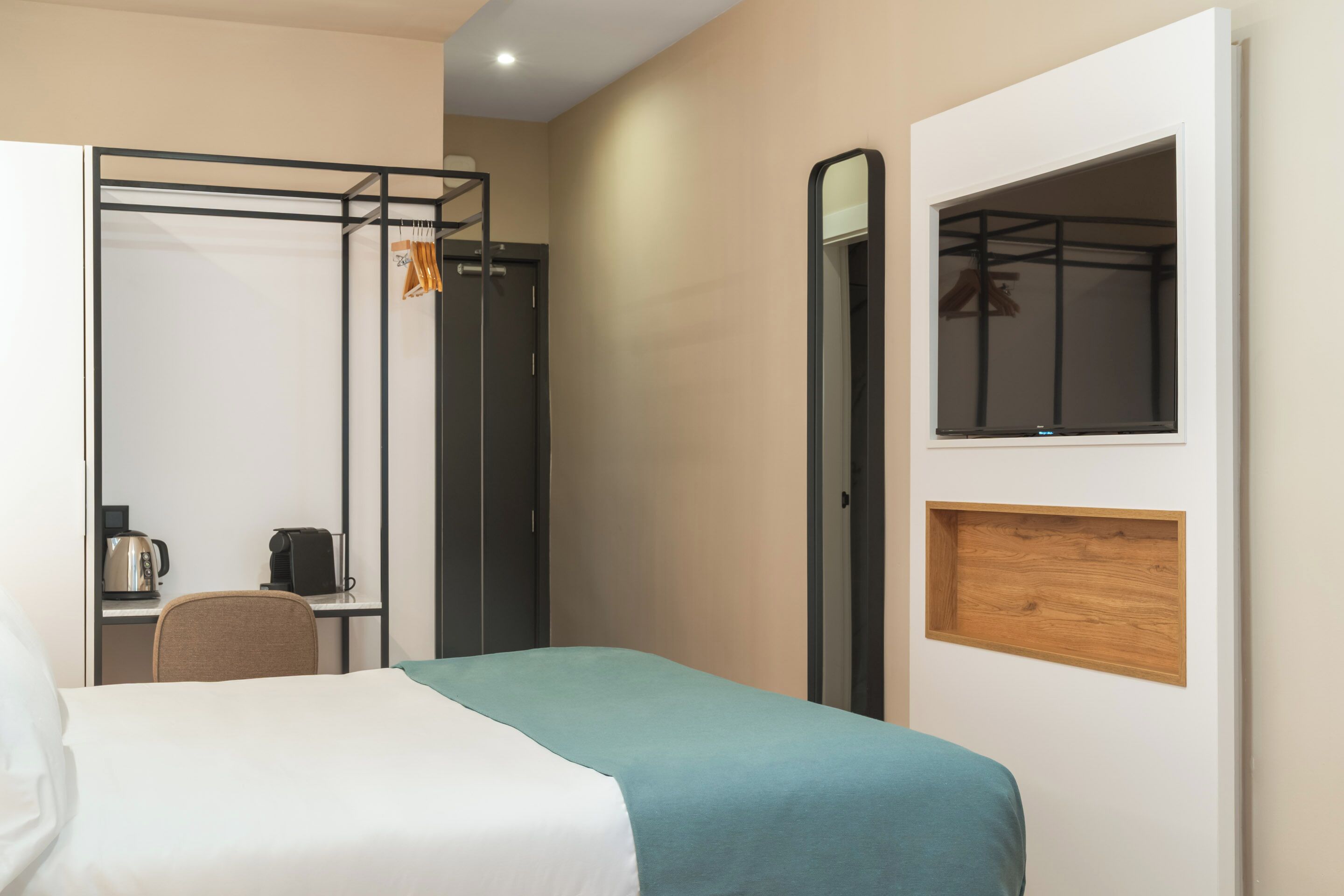 A hotel room with a bed, a desk with a kettle, a chair, and a wardrobe with a mirror.