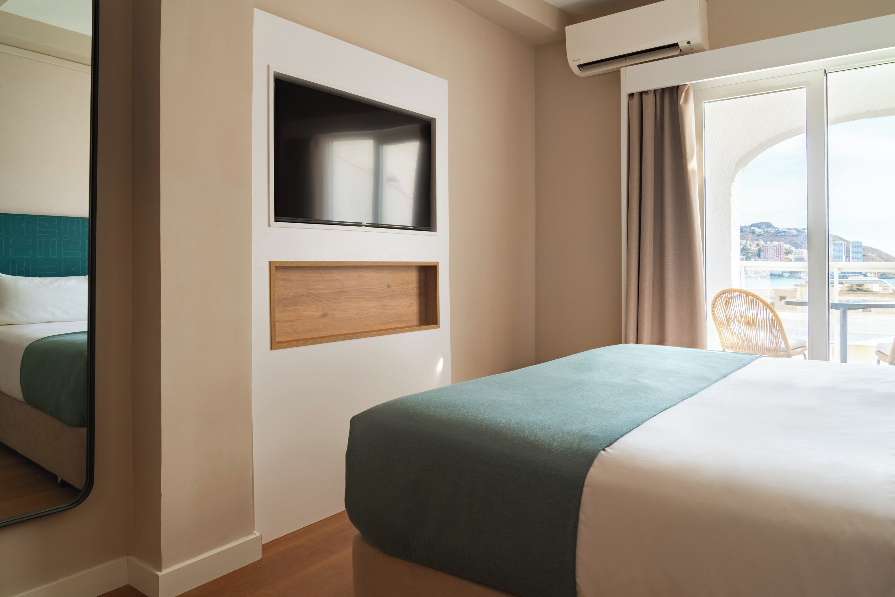 Habitación de hotel con cama, televisor montado en la pared, ventana con vistas y una silla cerca de la ventana.
