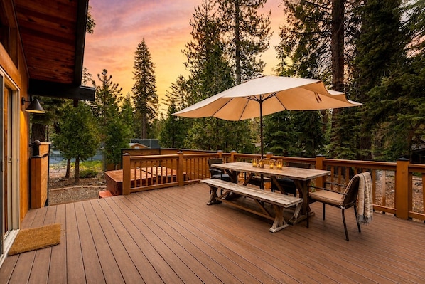 A wooden deck with a table and chairs, an umbrella, and a view of trees and a sunset.