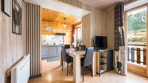Une pièce aux murs lambrés, avec une table à manger, des chaises, un téléviseur et, au fond, une cuisine.