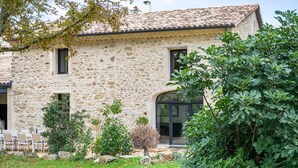 Un bâtiment en pierre avec un toit en tuiles, une porte d’entrée en verre et un petit patio équipé de chaises.