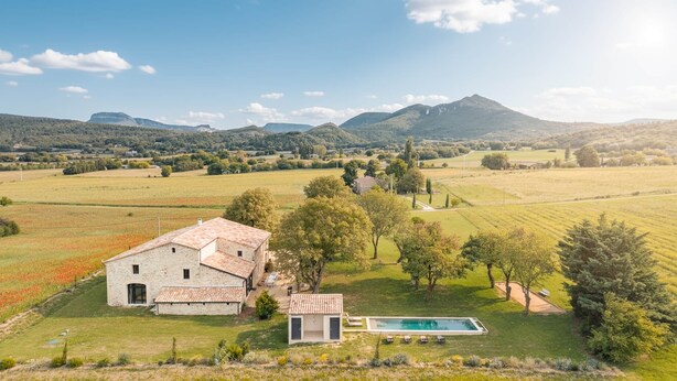 Une maison en pierre avec une piscine, entourée de champs et d’arbres, avec des montagnes en arrière-plan.