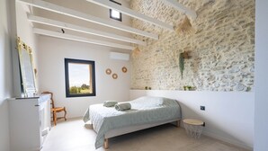 Une chambre avec un mur en pierre, un lit, une petite chaise en bois, une table ronde en bois et une fenêtre donnant sur un paysage verdoyant.