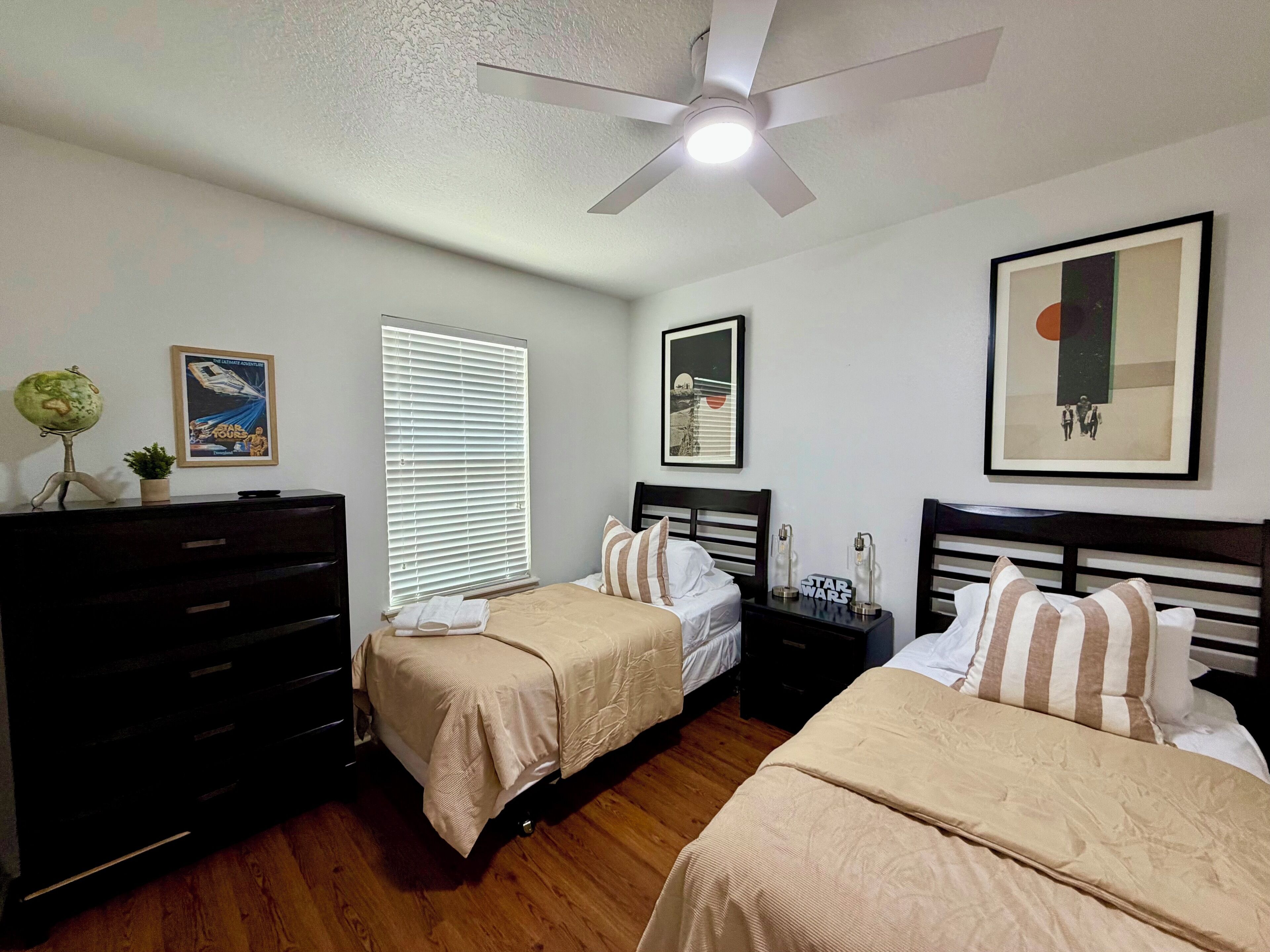 A bedroom with two beds, a dresser, a ceiling fan, and a window with blinds.