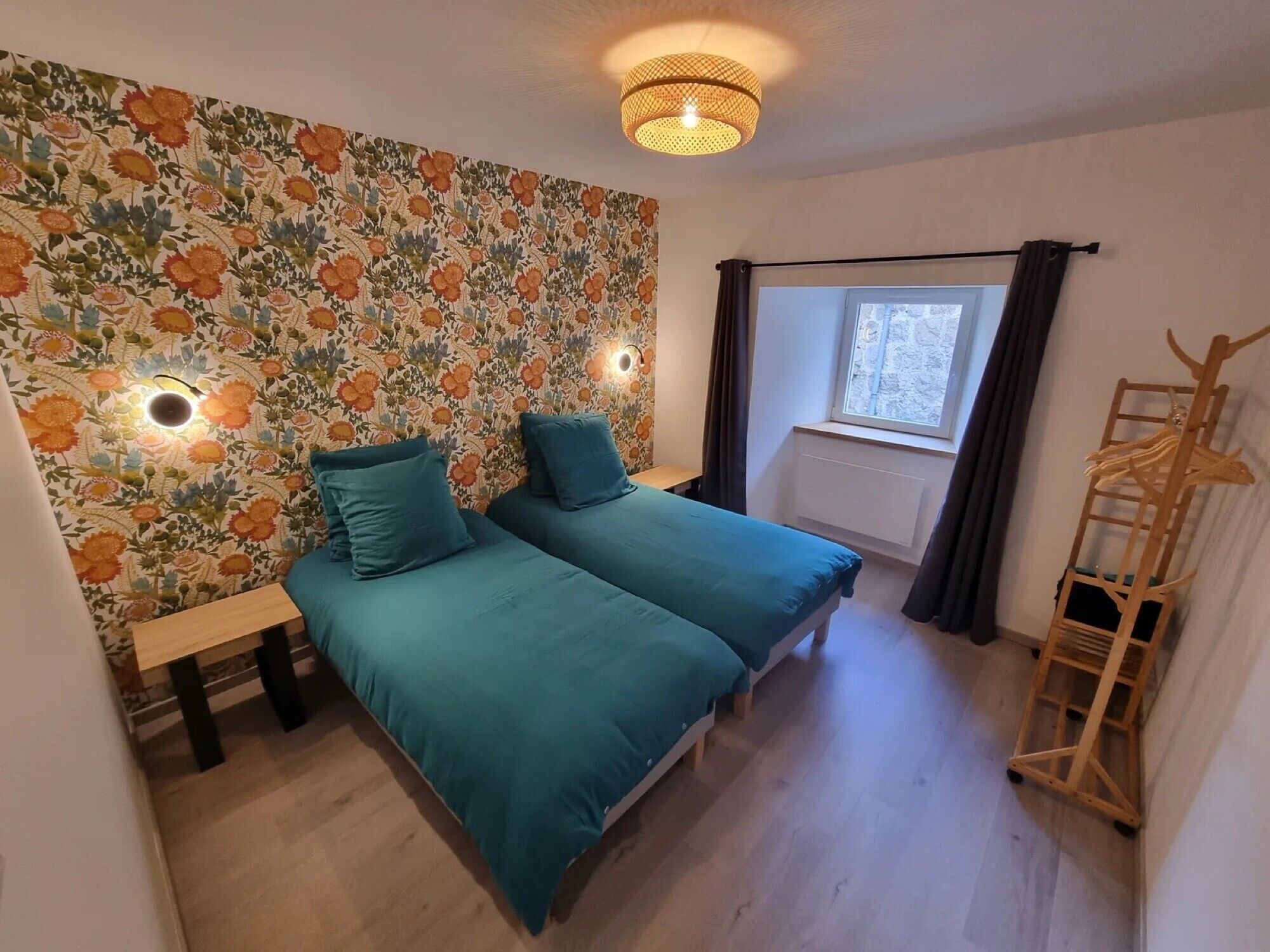 Quarto com duas camas, um banco de madeira, luminária fixada na parede, janela com cortinas e um cabideiro de madeira.
