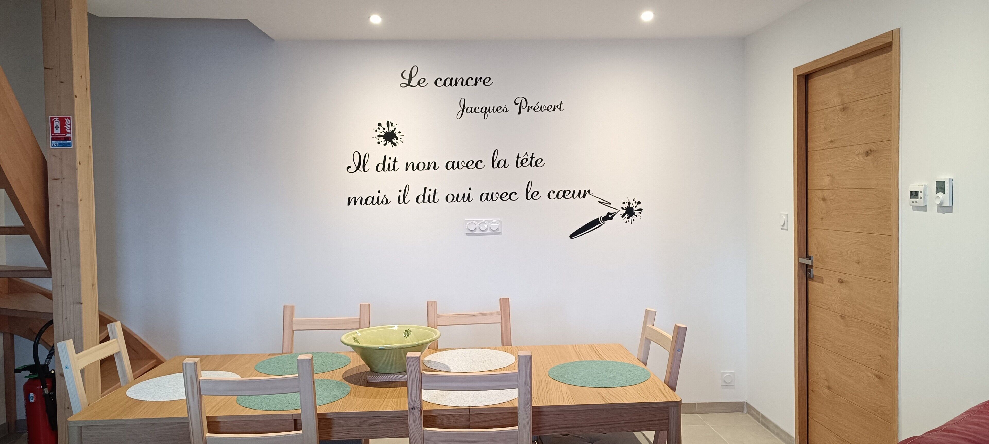 Un comedor con una mesa de madera, sillas y un tazón verde. Se aprecia un adhesivo en la pared con texto en francés y una ilustración a pluma.