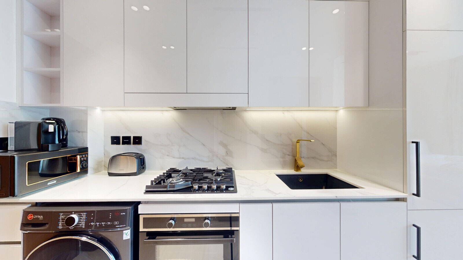 Una cucina moderna con mobili bianchi, forno a incasso, piano cottura a gas, lavello con rubinetto dorato e lavastoviglie.