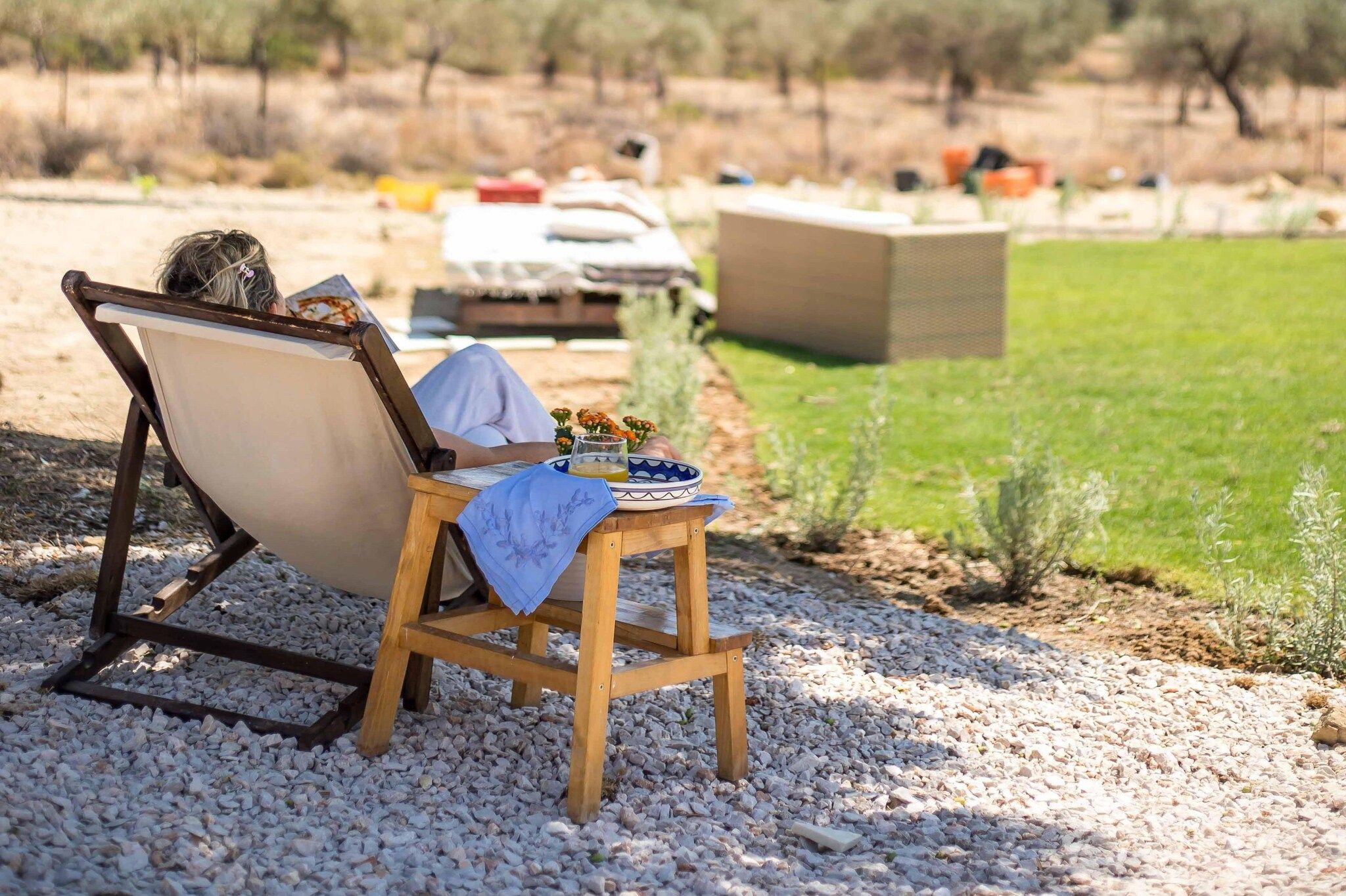Une personne est allongée dans un fauteuil, un livre à la main, un verre à portée de main et une assiette remplie de nourriture sur une table, le tout dans un cadre de jardin.