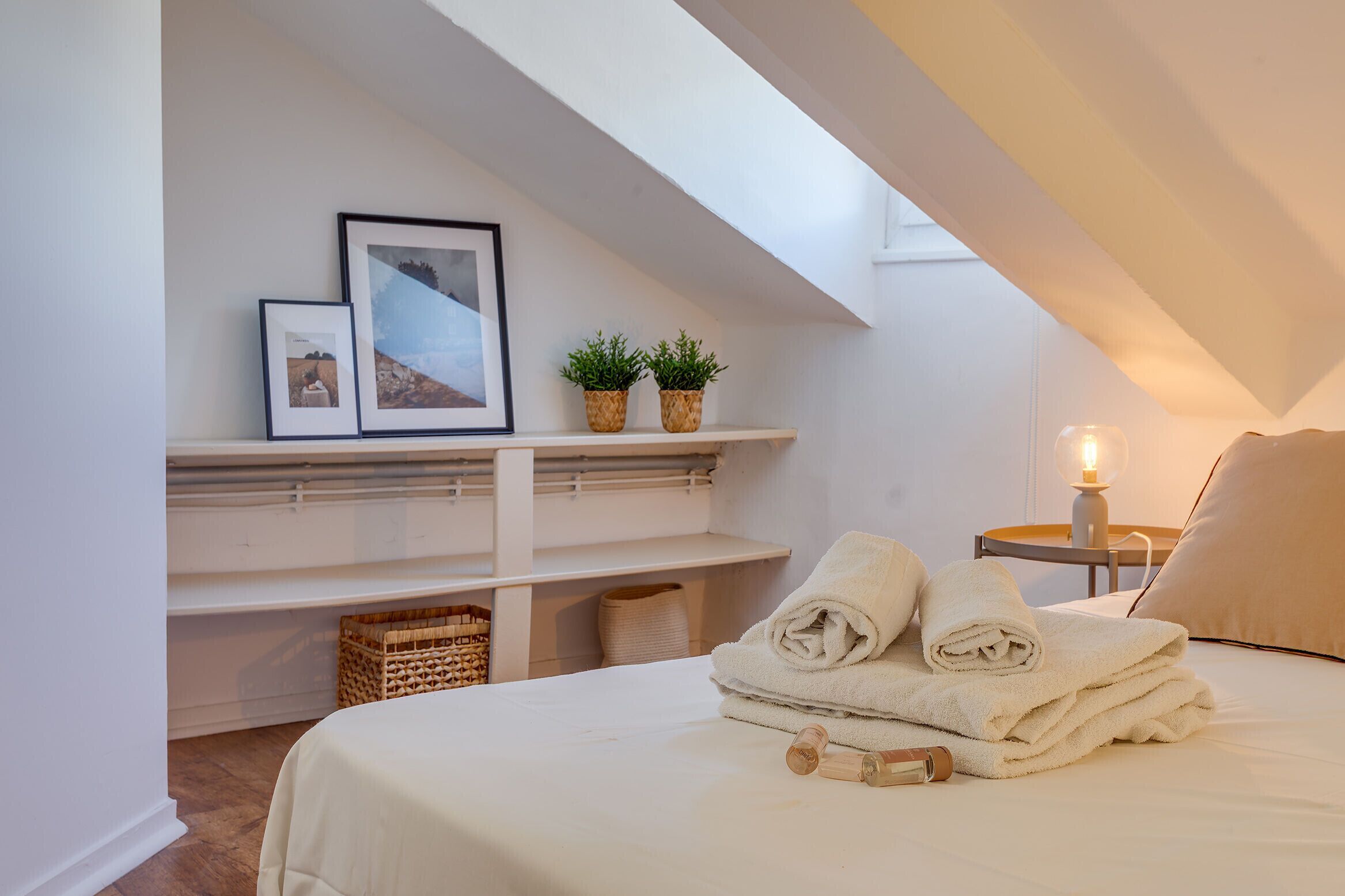 Een gezellige slaapkamer met een net opgemaakt bed, een klein nachtkastje met een lamp en een plank met ingelijste foto's en planten.