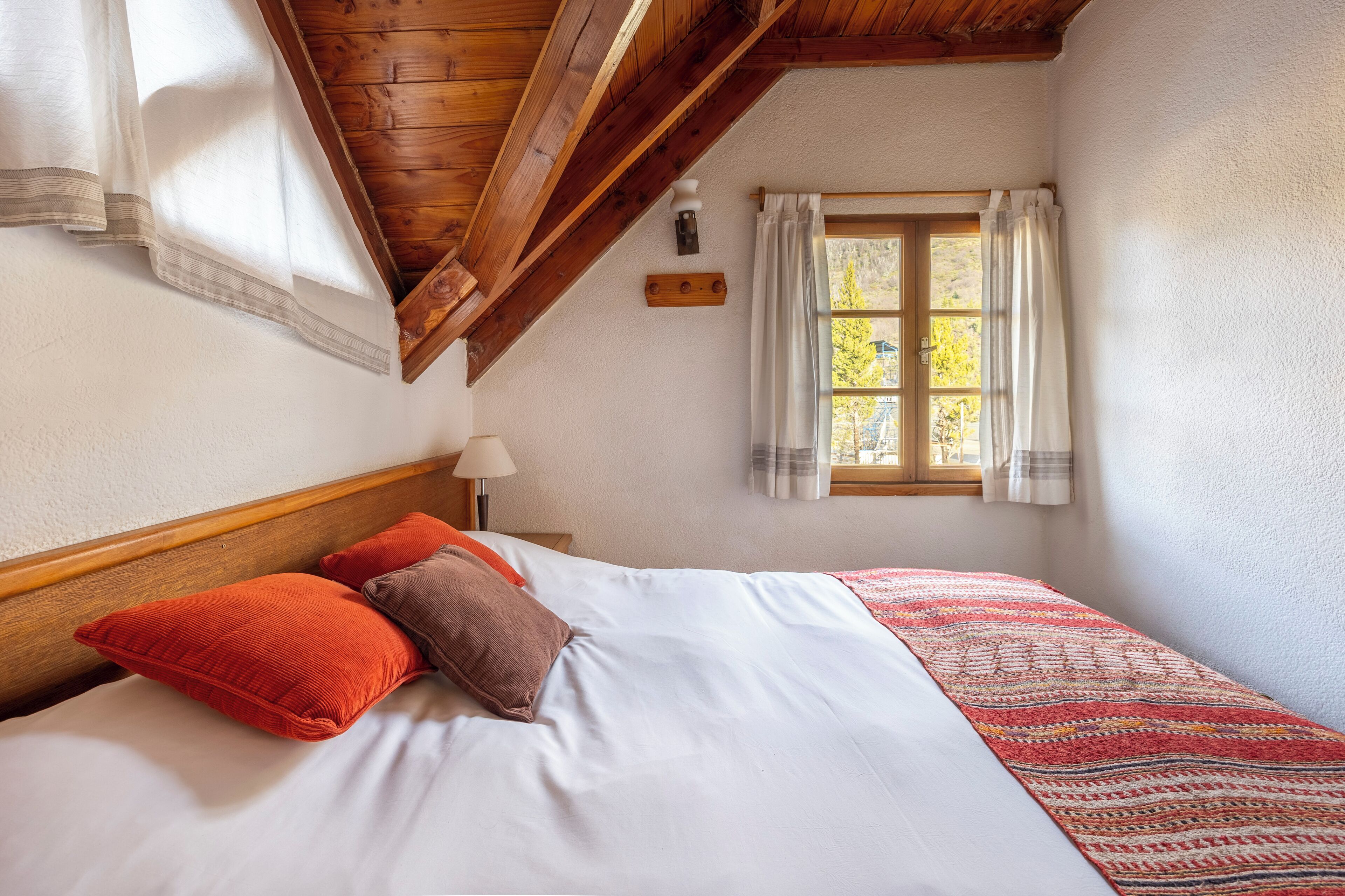 Un dormitorio con techo de madera, cama con ropa de cama blanca, dos almohadas, ventana con cortinas y vistas a árboles.