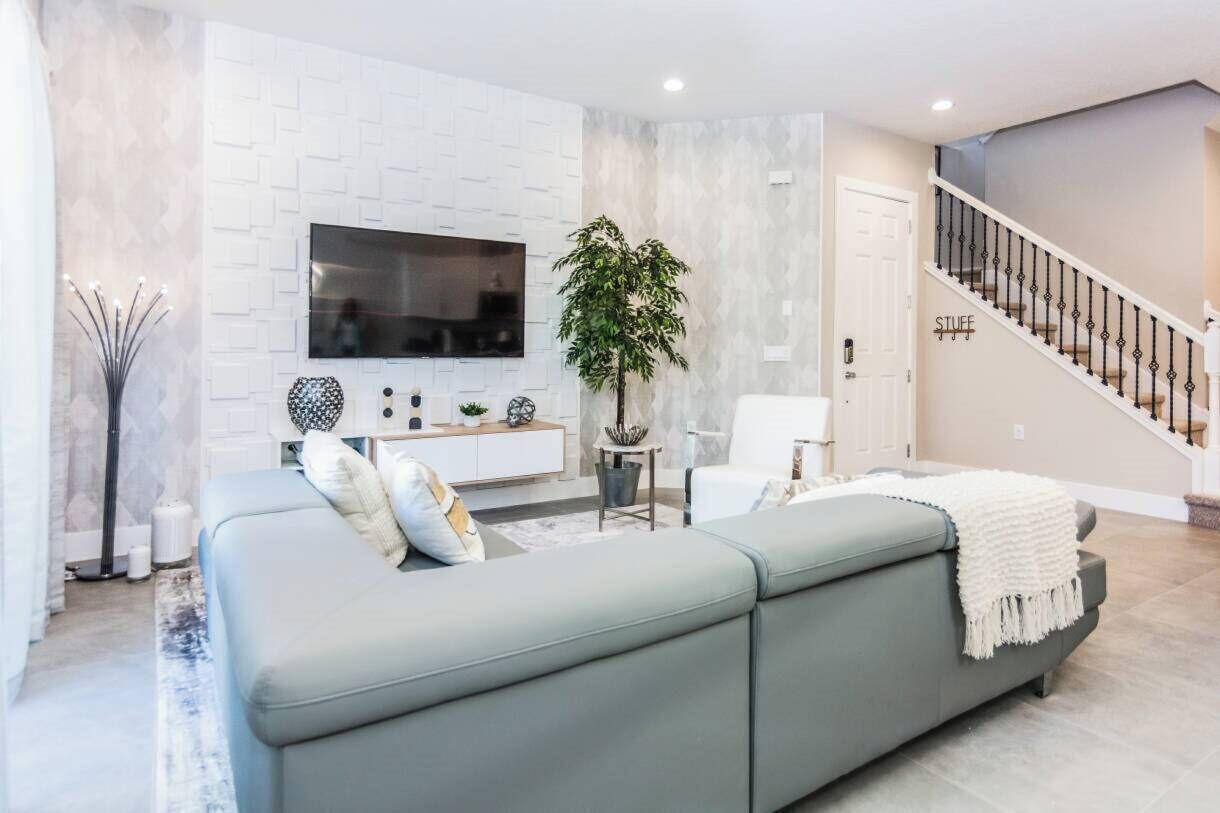 A modern living room with a grey sofa, a flat-screen TV mounted on a white brick-patterned wall, a staircase with a white railing, and a potted plant.