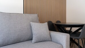 A grey sofa with a matching pillow and a black chair, positioned in front of a wooden slatted wall and a small round table.