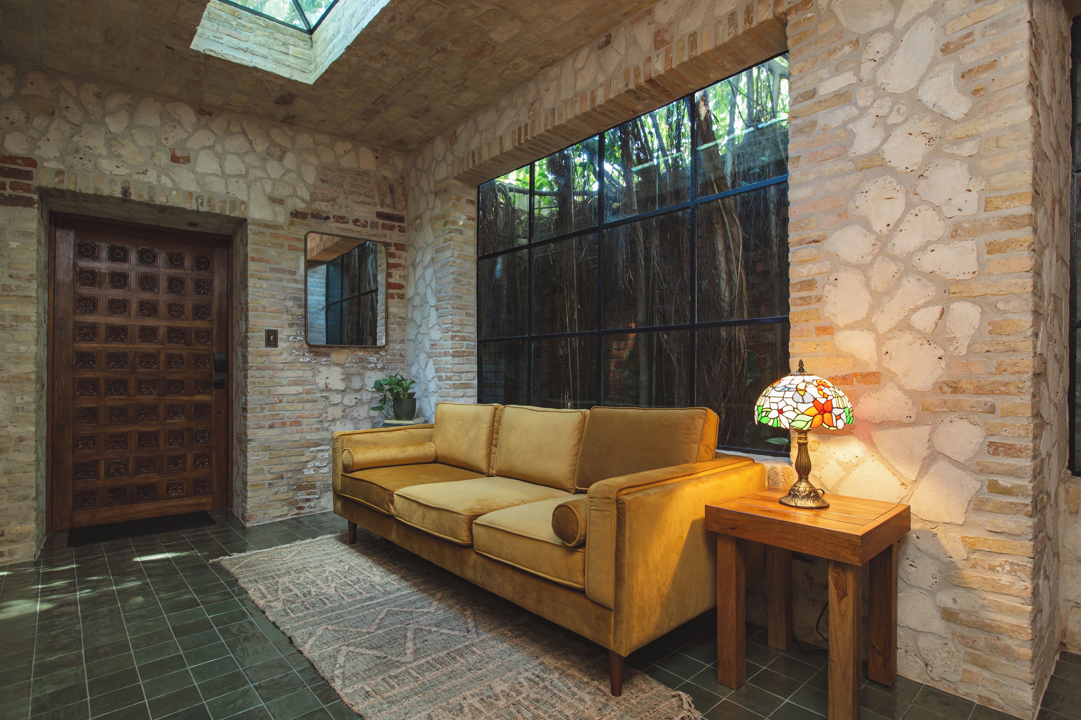 A room with a stone wall, a wooden door, a sofa, a table with a lamp, and a large window.