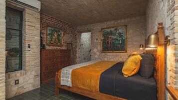 A room with a brick wall, a bed with a patterned bedspread, a wooden dresser, and two paintings.