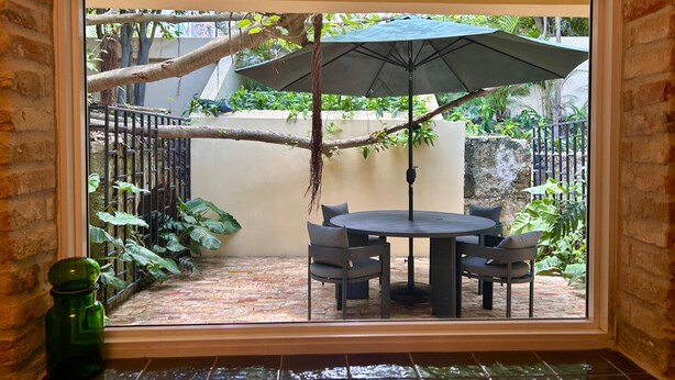 Een terras met een ronde tafel en stoelen, een parasol en uitzicht op een tuin door een raam.