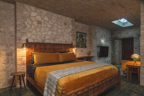 Un dormitorio con pared de piedra, cama de madera con cabecera, mesita de noche, lámpara y tragaluz.