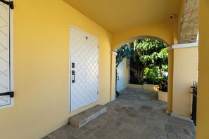 A corridor with a white door marked with a 'B', leading to a covered area with a brick wall and greenery. - Treehouse - The Nest (Christiansted)