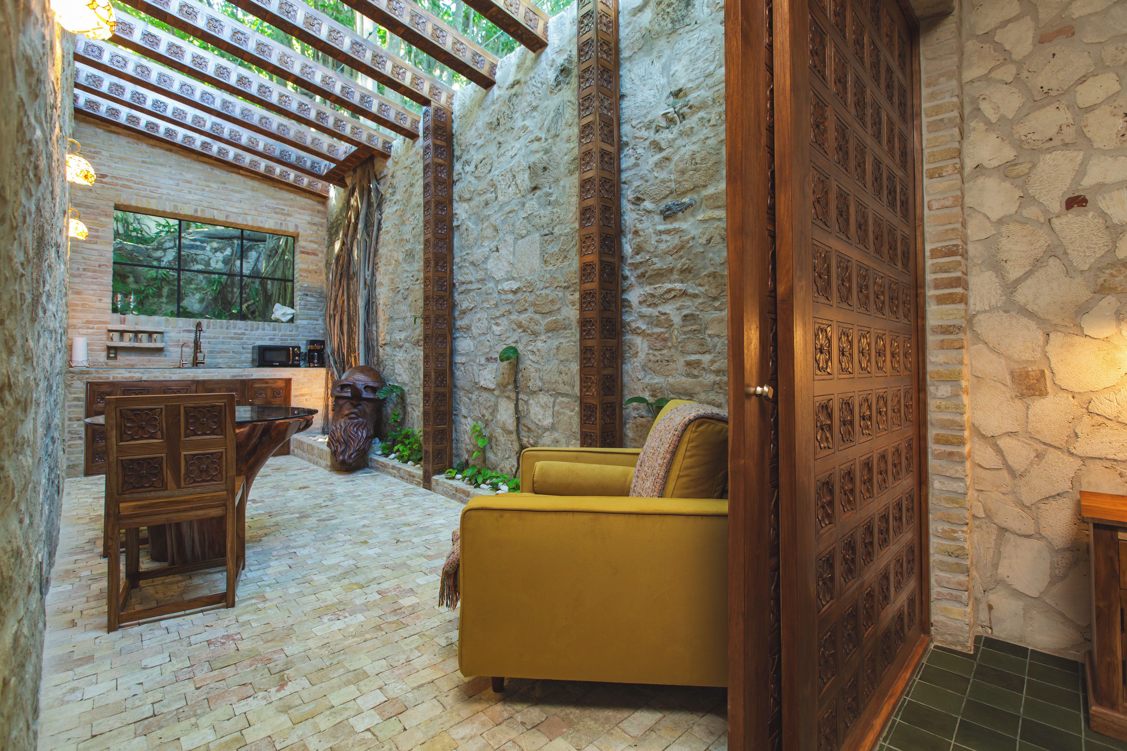 A cozy interior with a stone wall, a wooden door with intricate carvings, a yellow sofa, and a brick floor.