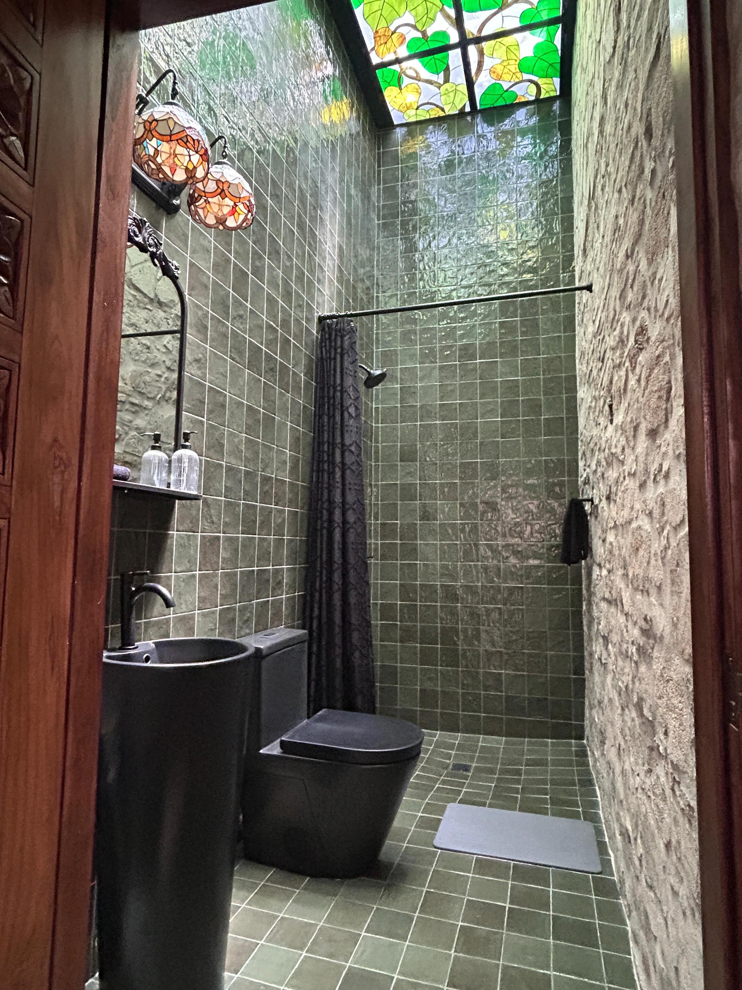 A bathroom with a sink, toilet, and shower area, featuring green tiled walls and a stained glass window.