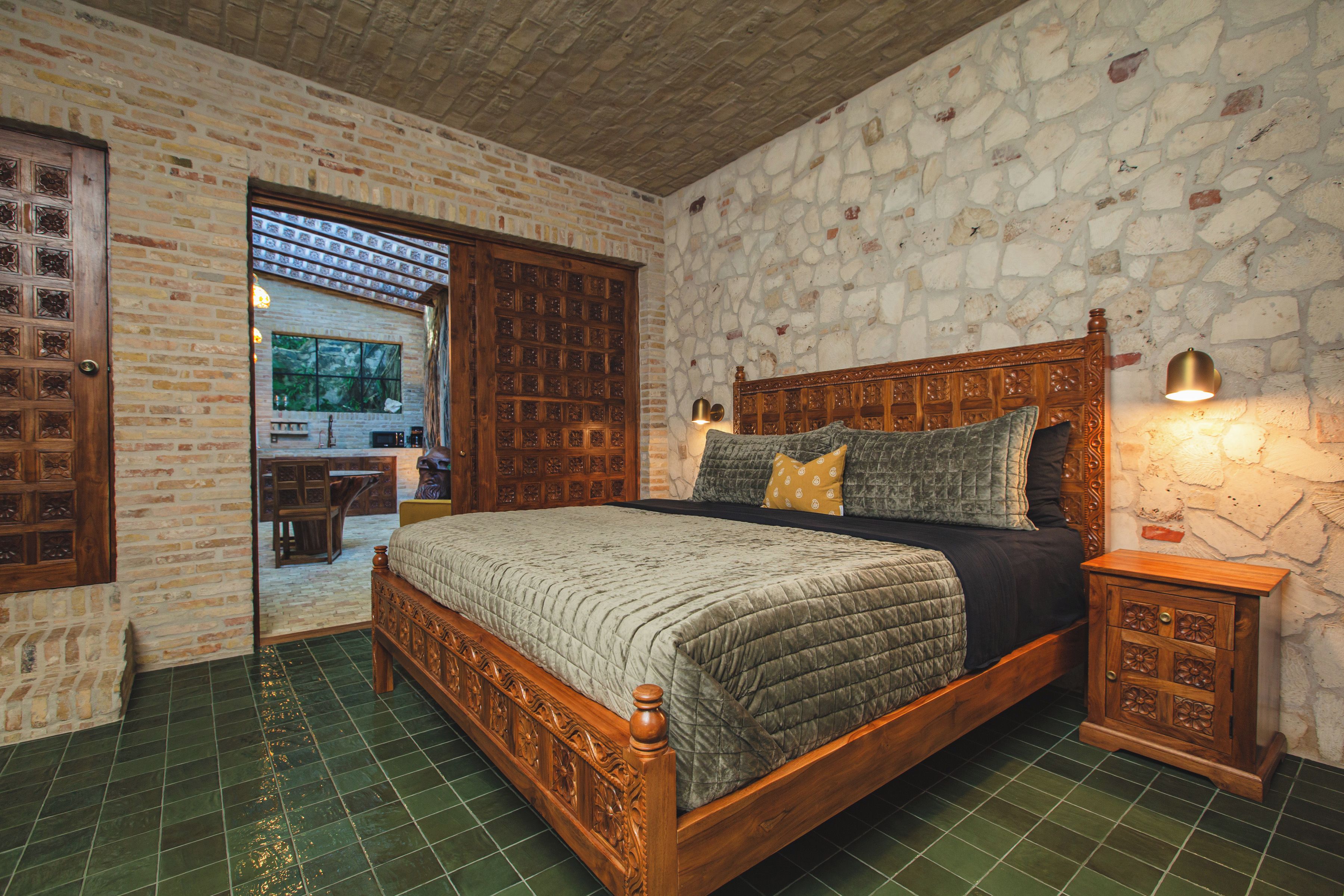 A bedroom with a large bed, a wooden headboard, a nightstand, and a stone wall.