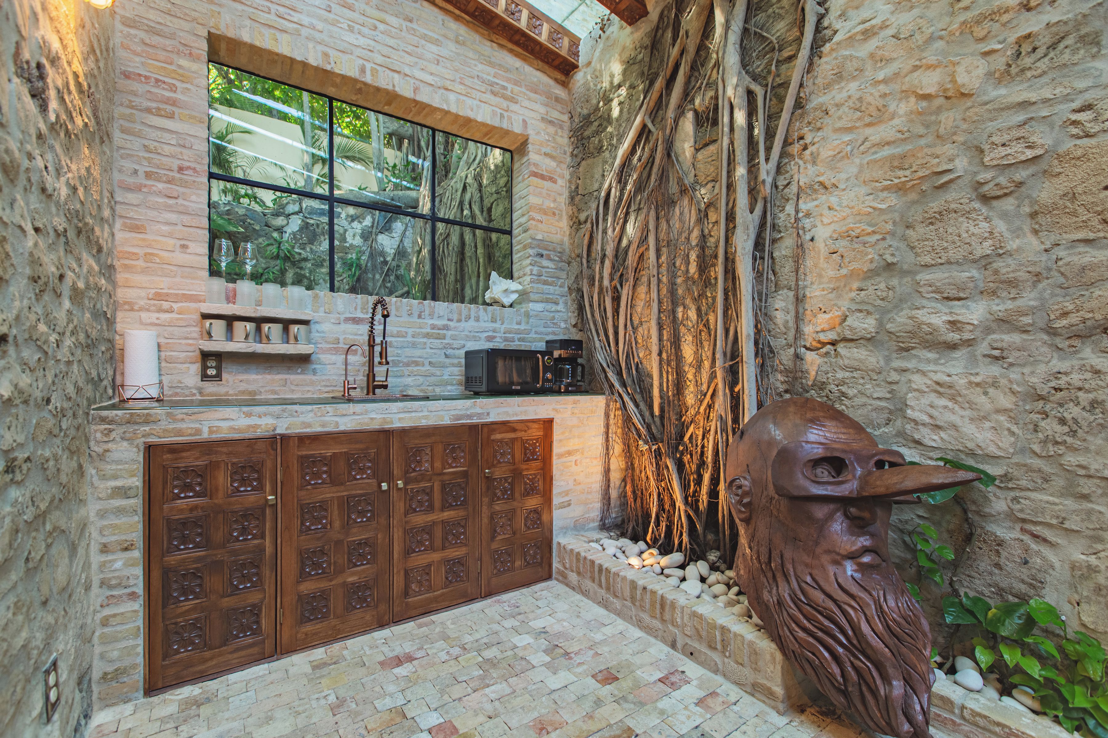 A rustic kitchen with a brick wall, wooden cabinets, and a window overlooking greenery.