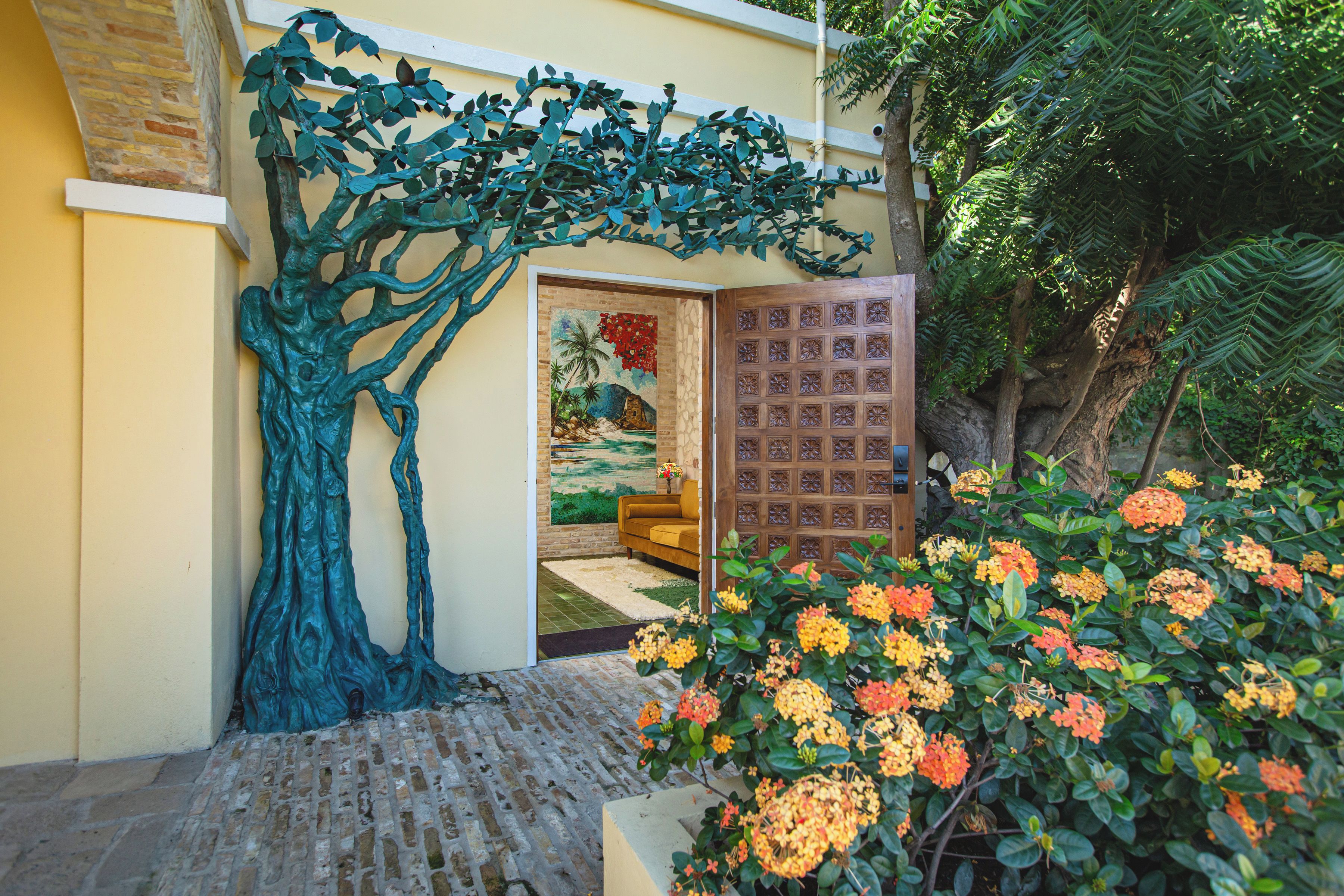 A doorway with a tree sculpture, a wooden door with floral carvings, and a garden with flowering plants.