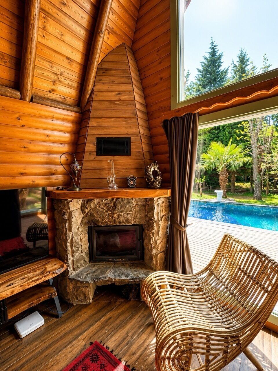 Interior de una cabaña de madera con chimenea de piedra, un sillón de mimbre y vista a una piscina.