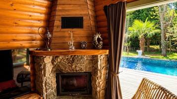 Interior de una cabaña de madera con chimenea de piedra, un sillón de mimbre y vista a una piscina.
