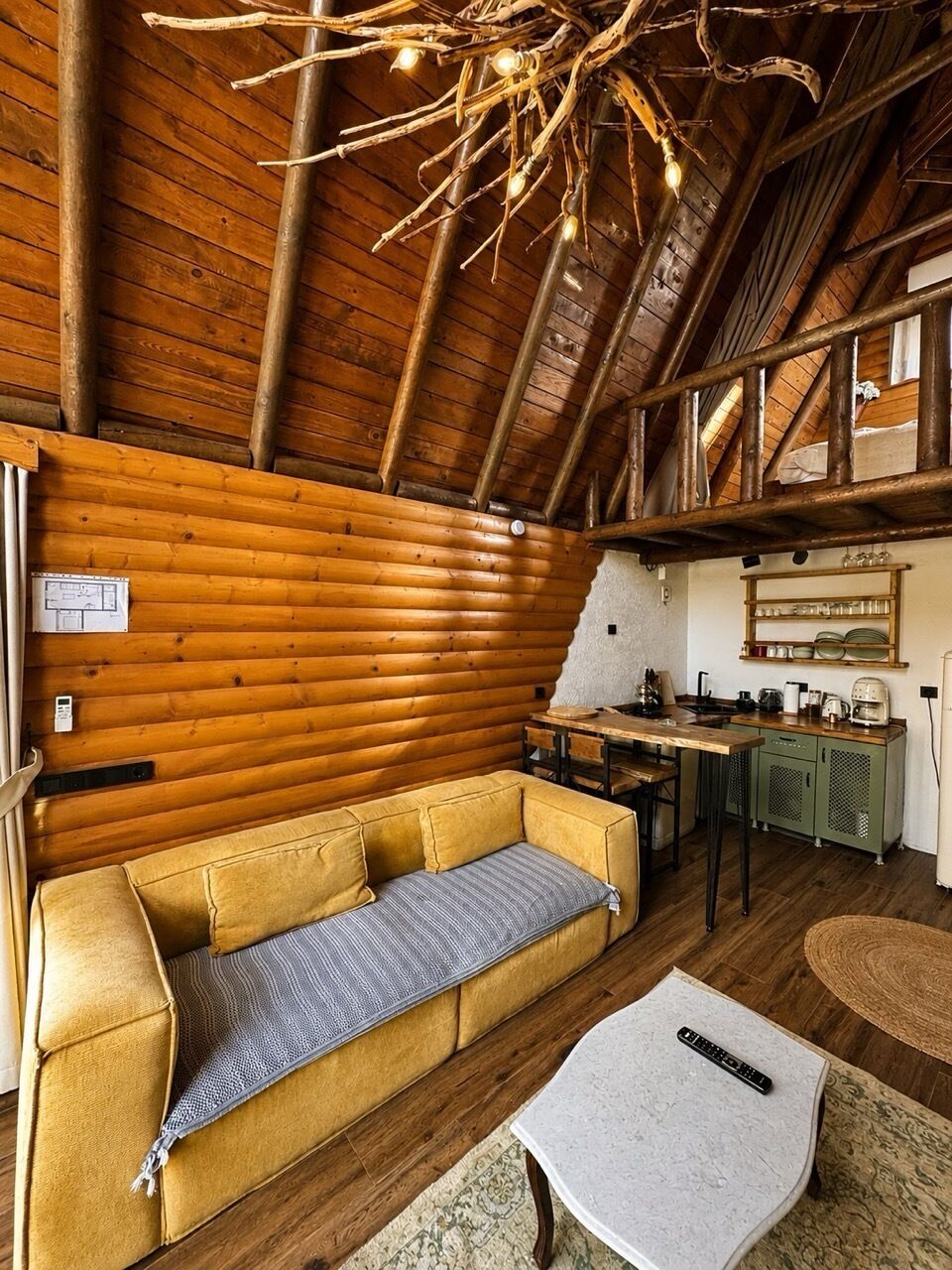 A log cabin interior with a wooden ceiling, a sofa, a small table, and a kitchen area.