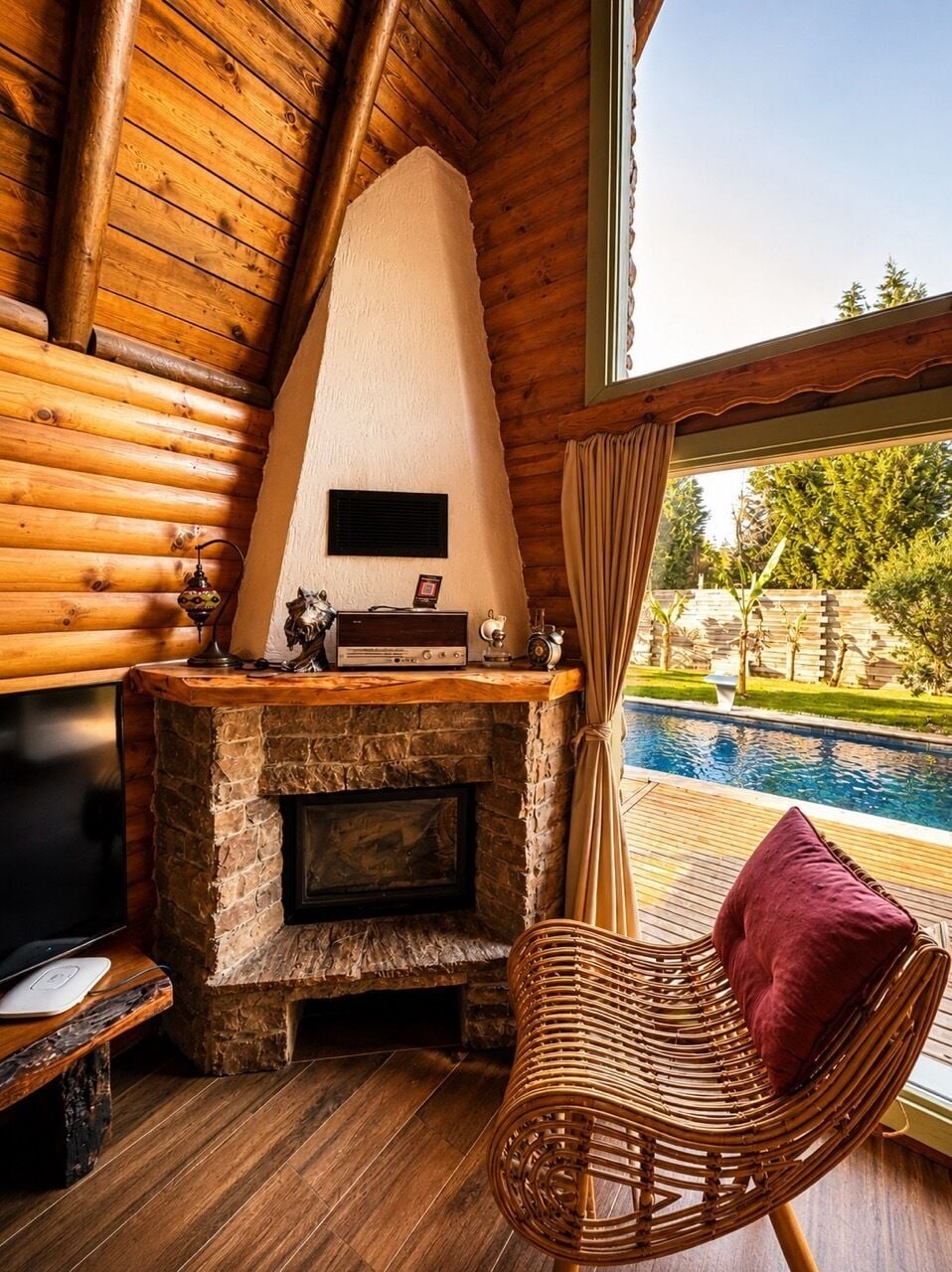 Ein gemütliches Blockhaus-Interieur mit Kamin aus Stein, einem Korbsessel und Blick auf einen Swimmingpool.