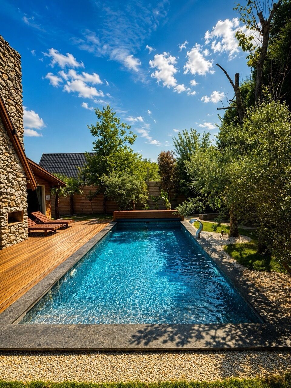 A rectangular swimming pool with a wooden deck and lounge chairs, surrounded by greenery and a stone building.