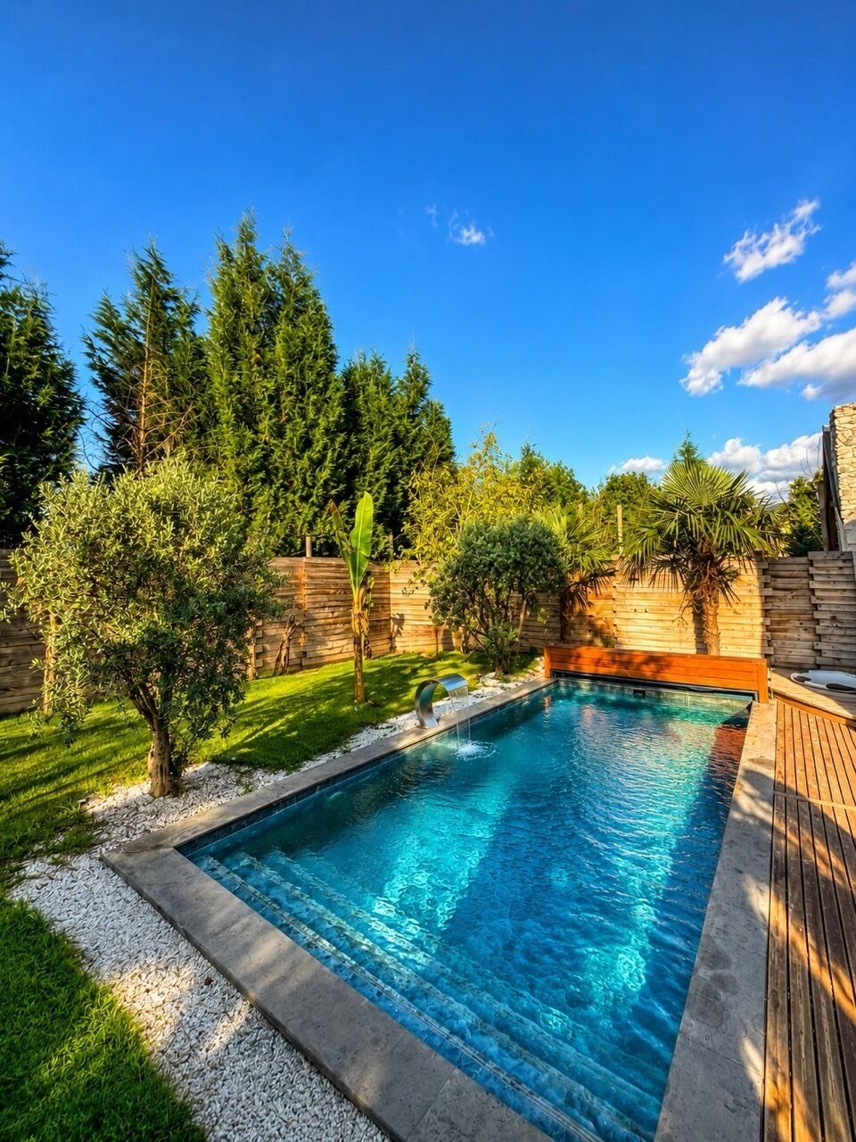 A rectangular swimming pool with a wooden deck surrounding it, set against a backdrop of trees and a clear sky.
