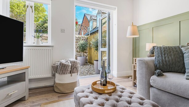 A living room with a television, a grey sofa, a wicker ottoman, and a glass door leading to an outdoor area with plants and a brick wall. - BRAND NEW! Renovated luxury 2-bed cottage, parking & garden (Lechlade)
