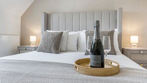 A neatly made bed with a bottle of wine and a glass on a woven tray, set against a modern bedroom with a sloped ceiling and a wooden headboard. - BRAND NEW! Renovated luxury 2-bed cottage, parking & garden (Lechlade)