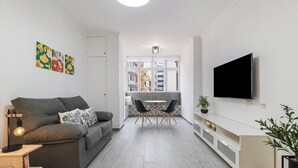 A modern living room with a grey sofa, a dining table with chairs, a flat-screen TV mounted on the wall, and a small side table with a lamp.