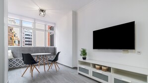 A modern living room with a large TV, a dining area with chairs and a table, and a window with a city view.
