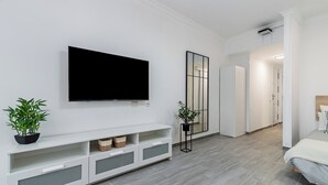 A modern hotel room with a flat-screen TV mounted on the wall, a white console table with storage, a large mirror, and a wooden floor.