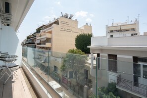 Un balcon doté d’une balustrade en verre, donnant sur un quartier résidentiel avec des immeubles et un arbre.