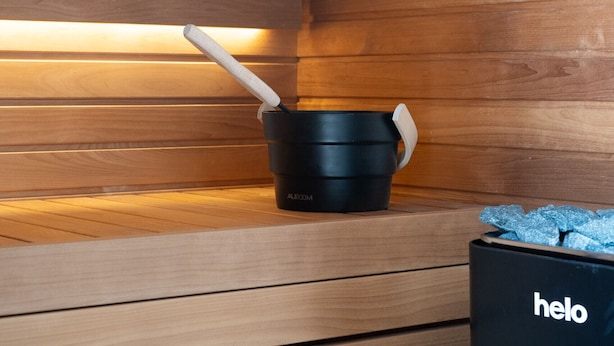 A sauna room with wooden walls and benches, a black bucket, and a black bag labeled 'helo'.