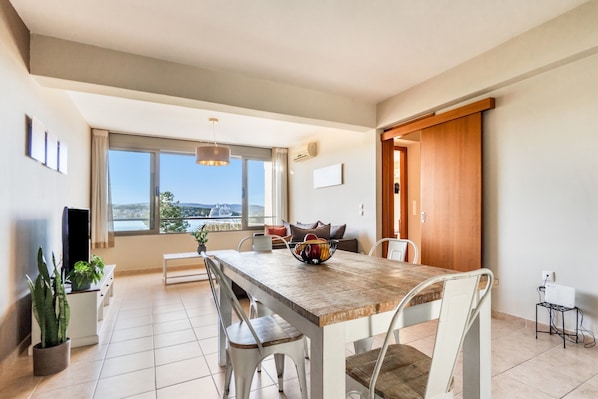 A modern dining area with a wooden table, white chairs, a bowl of fruit, and a view of the outdoors.