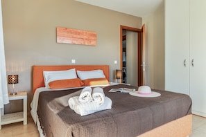 A neatly made bed with a brown bedspread, a white pillow, and a rolled white towel. There is a white hat on the bed, and a wooden headboard.