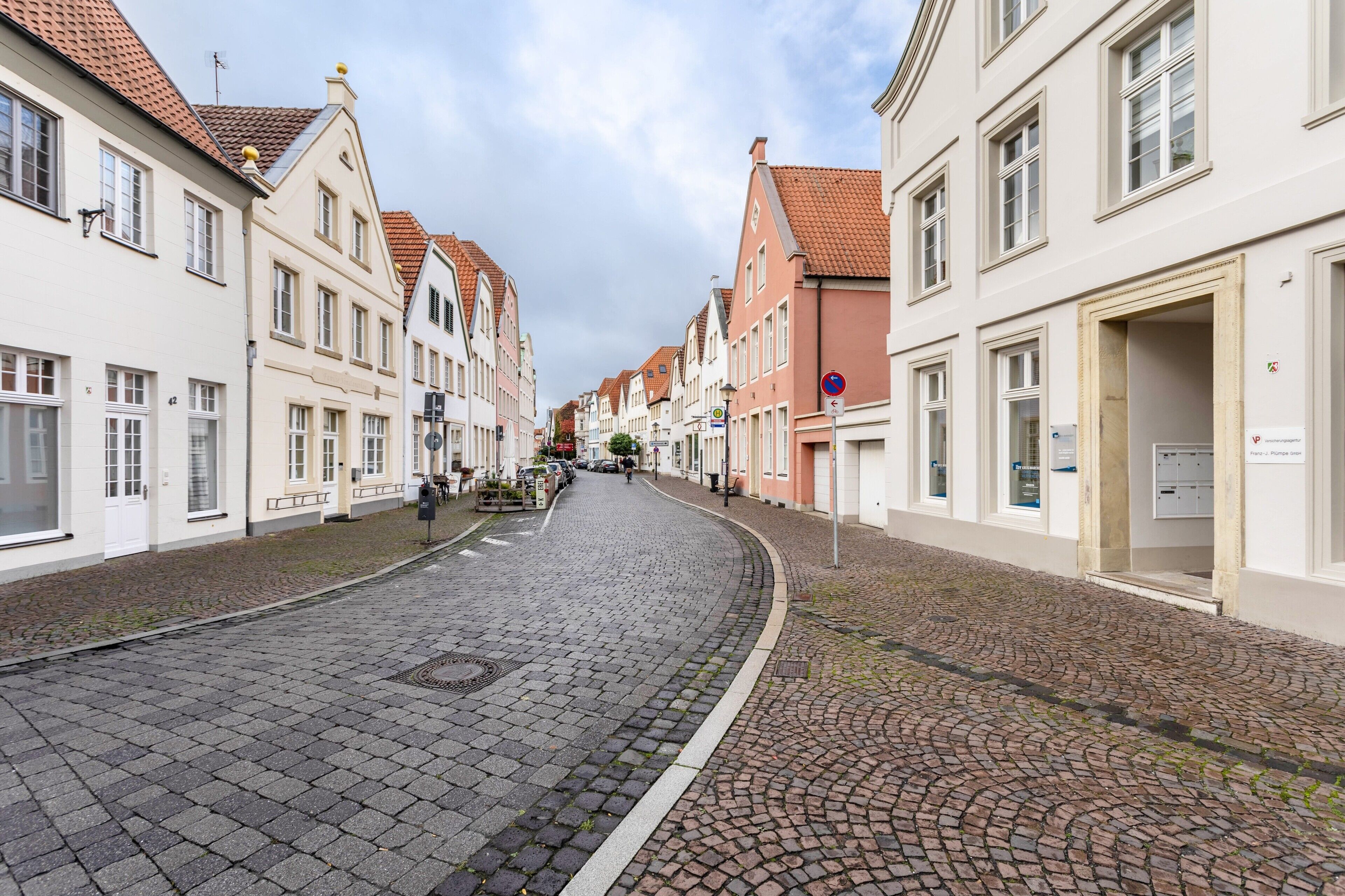 Een straat met keien, geflankeerd door historische gebouwen, een bord dat parkeren verbiedt, en een heldere lucht.