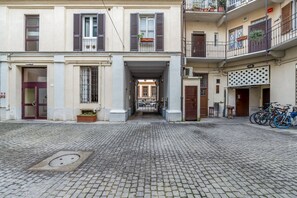 A cobblestone street leading to a building with a central entrance, balconies with plants, and bicycles parked outside.