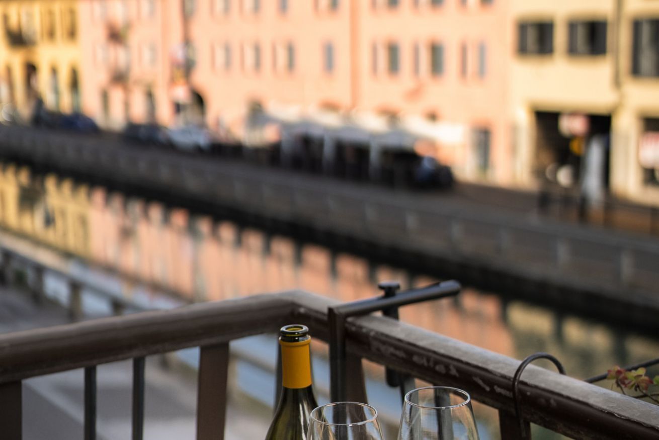 Una botella de vino y dos copas sobre una barandilla de balcón con vistas a un canal y edificios al fondo.